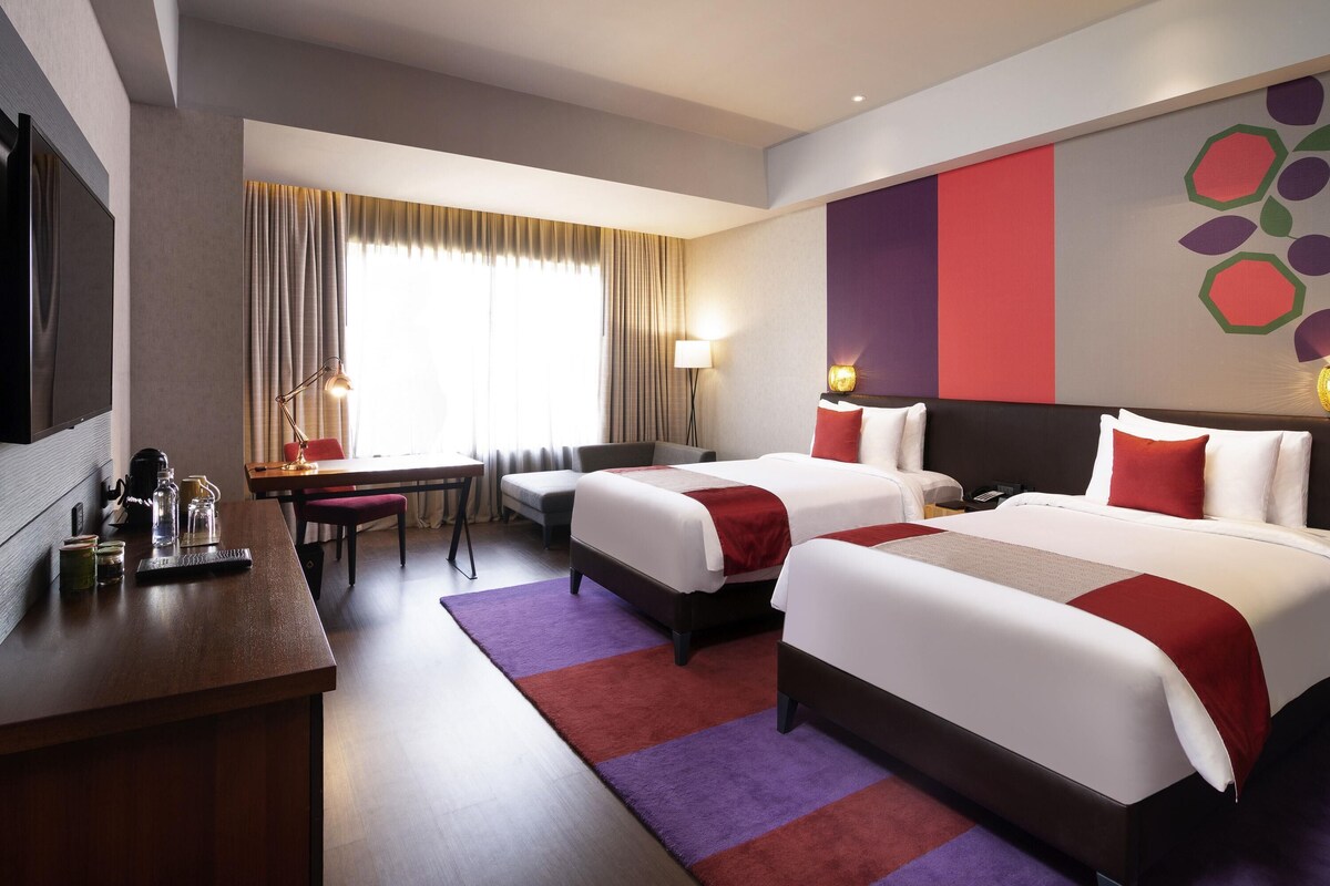 A hotel room with two beds, a desk, a chair, a TV, and a large window with curtains.