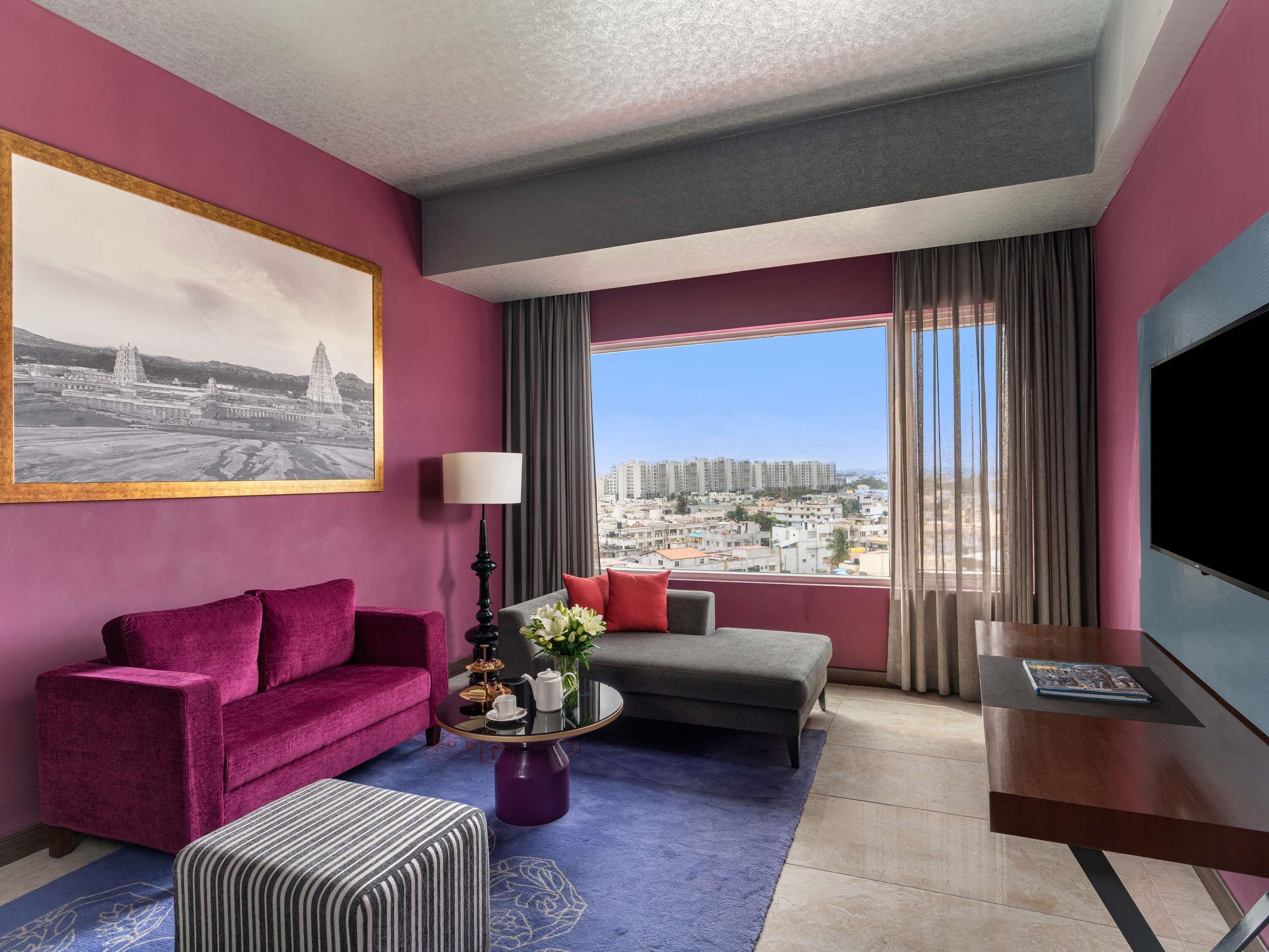 A hotel room with a purple sofa, a grey armchair, a striped ottoman, a coffee table with flowers, a TV, and a large window with a city view.