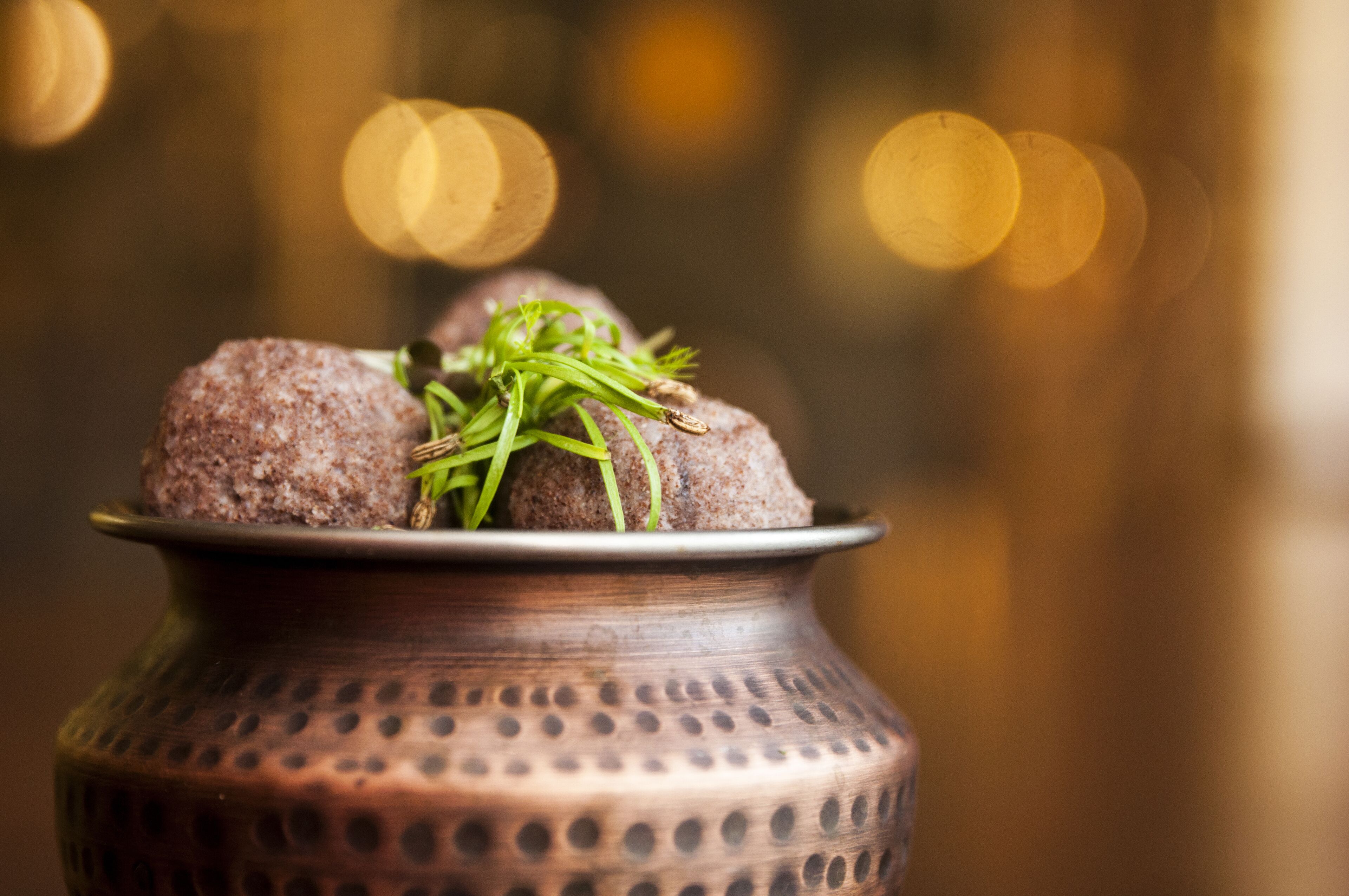 A traditional copper pot with a plate of food, possibly a Middle Eastern dish, with a blurred background featuring warm bokeh lights.