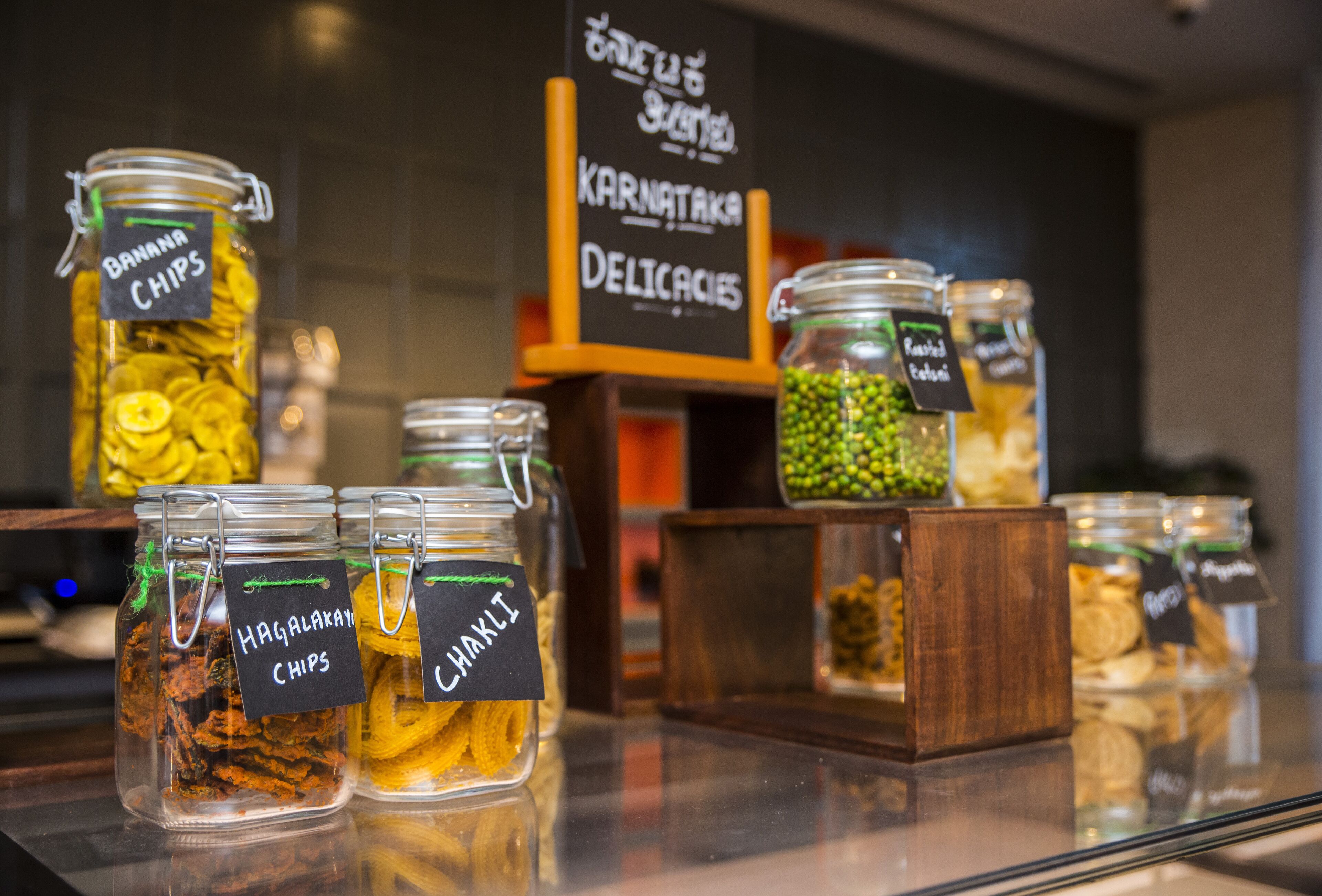 A display of various snacks in glass jars with labels, including banana chips and chips, in a store setting.