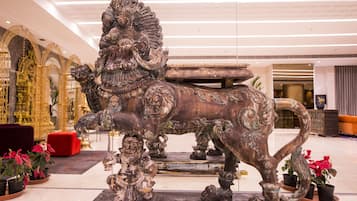 Une grande statue de bronze représentant un lion, ornée de motifs complexes, est placée sur un piédestal dans un vaste hall d’entrée à l’architecture moderne, où se trouve également un banc rouge.