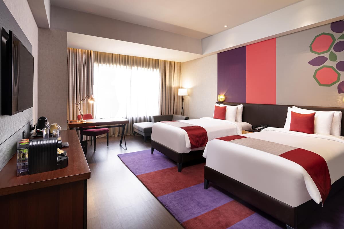Ein Hotelzimmer mit zwei Betten, einem Schreibtisch mit Kaffeemaschine, einem Fernseher und einem großen Fenster mit Vorhängen.
