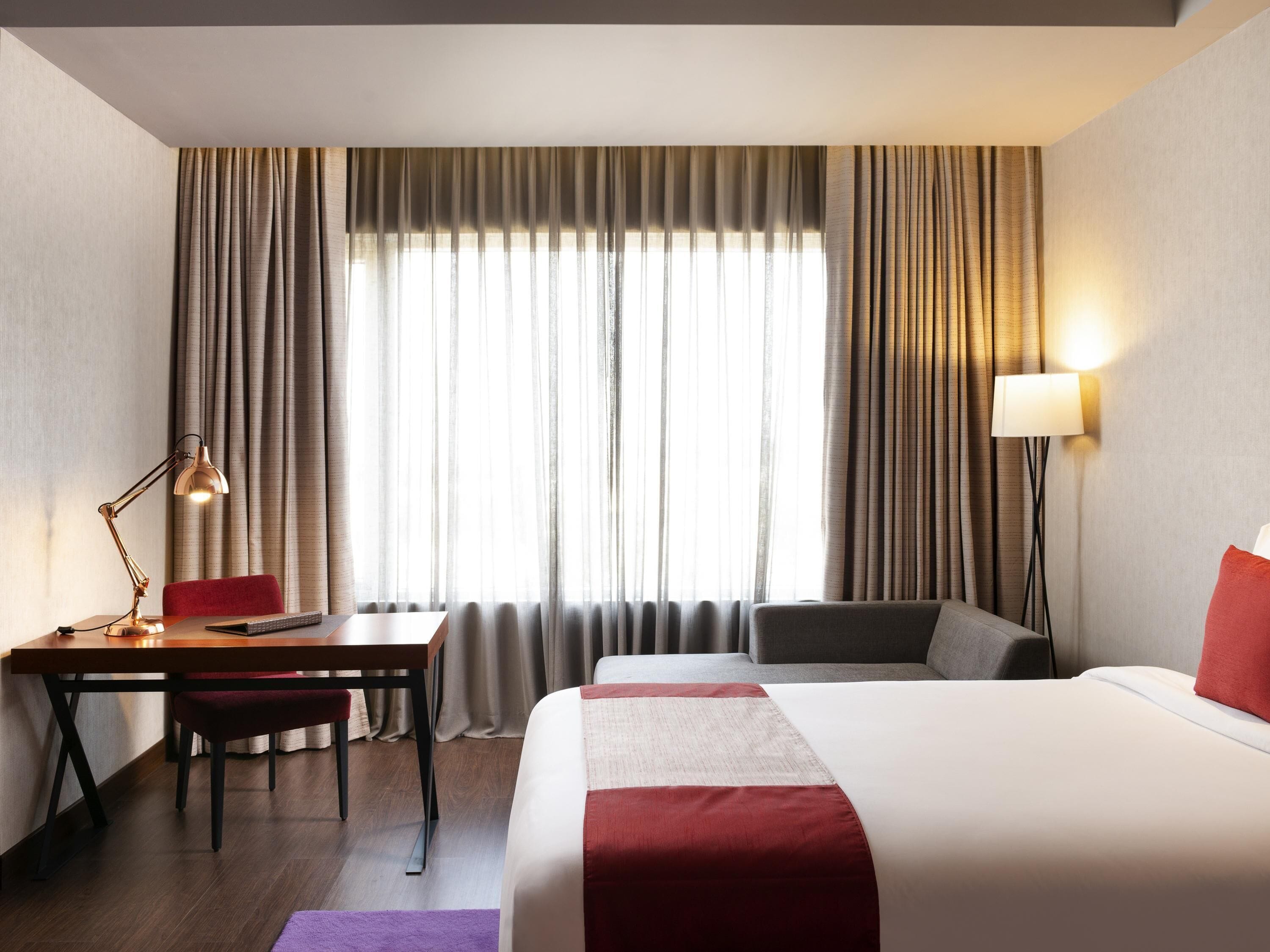 Ein Hotelzimmer mit Bett, Schreibtisch mit Stuhl, Lampe und Blick durch Vorhänge nach draußen.