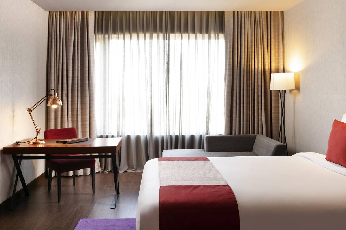 Habitación de hotel con cama, escritorio con silla, lámpara y vistas al exterior a través de cortinas.