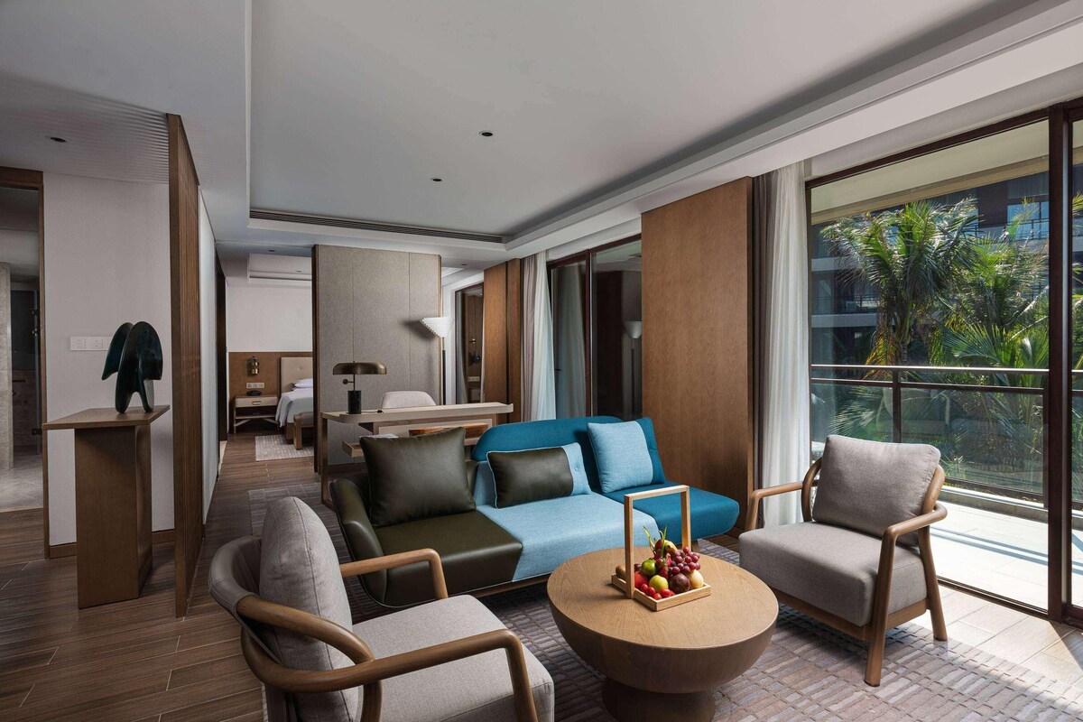 Una habitación de hotel moderna con sofá, sillas, una mesita de centro y un amplio ventanal con vistas a palmeras.