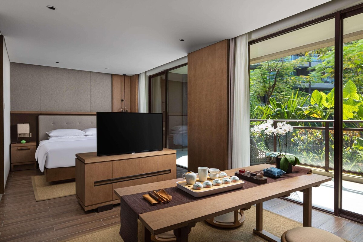 Una habitación de hotel moderna con una cama, un televisor, una mesa de comedor y una bandeja de comida, además de un ventanal con vistas a zonas verdes.