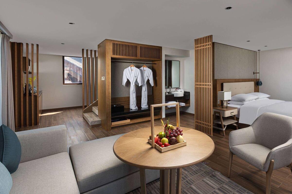 A hotel room with a bed, a desk, a chair, a sofa, a wardrobe, and a table with fruit.