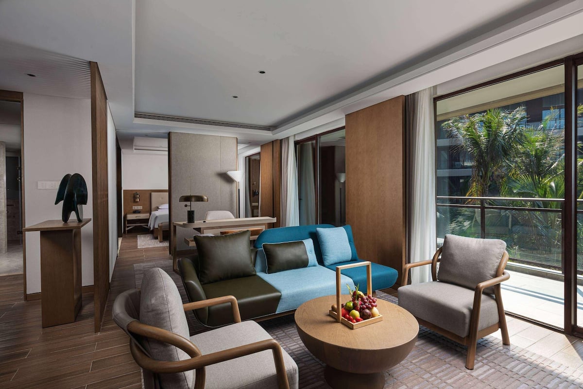 A modern hotel room with a sofa, chairs, a coffee table, and a large window with a view of palm trees.