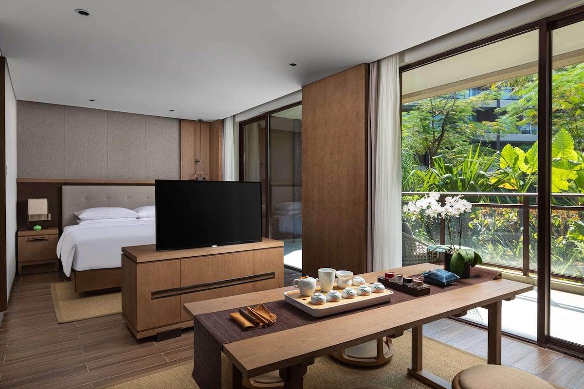 A modern hotel room with a bed, a TV, a dining table with a tray of food, and a large window with a view of greenery.