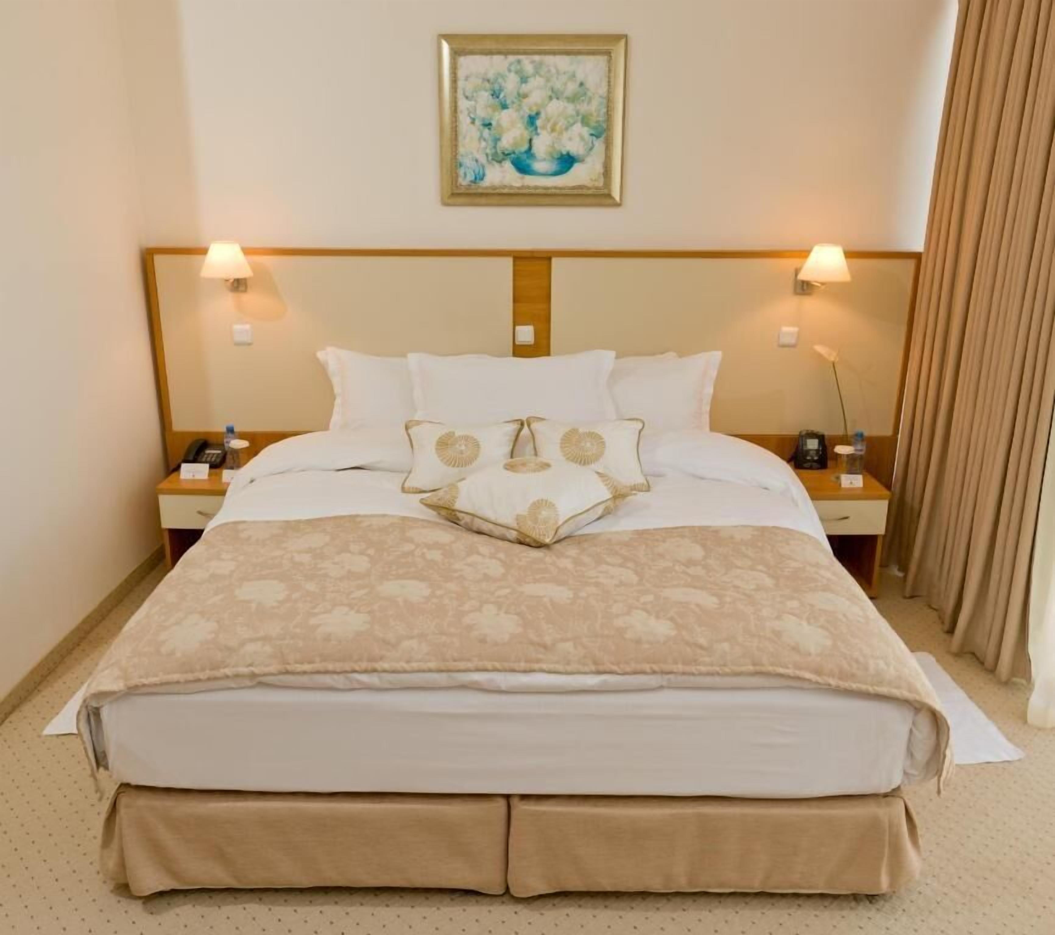 Une chambre d’hôtel avec un grand lit, deux tables de chevet avec des lampes, un tableau au-dessus du lit et un rideau sur le côté droit.