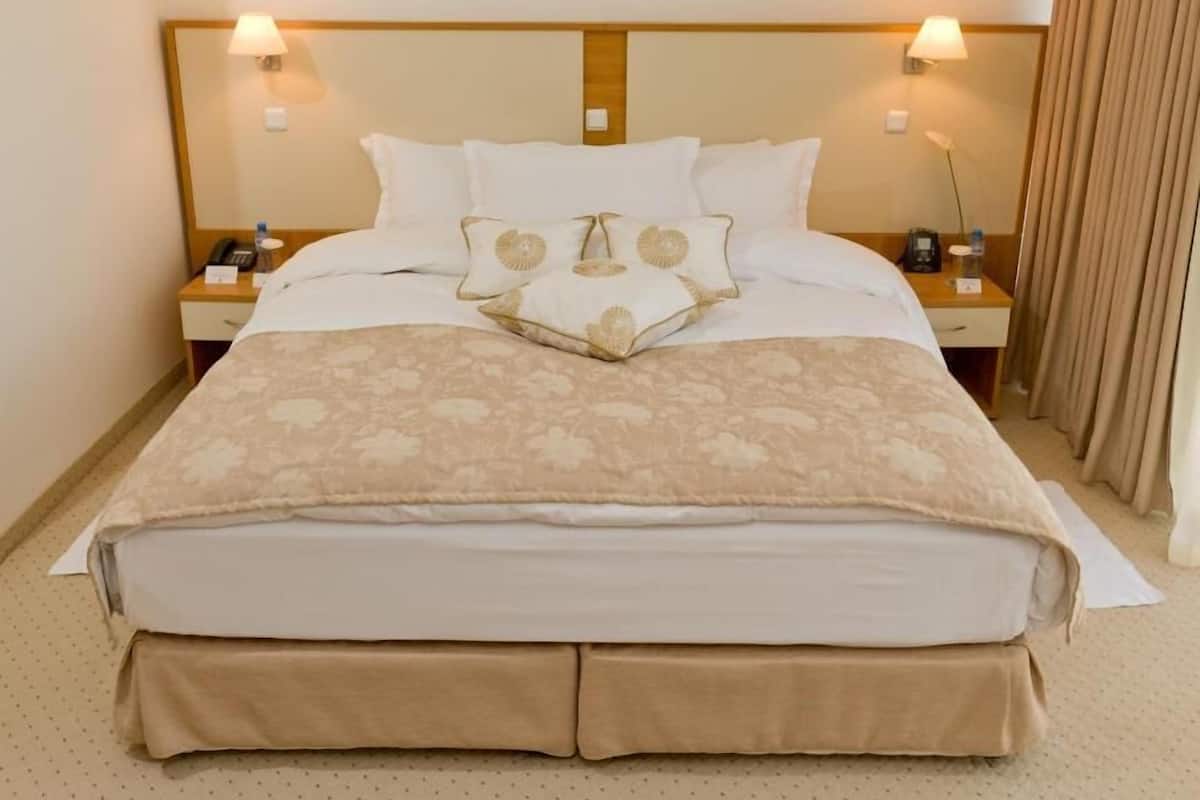 Une chambre d’hôtel avec un grand lit, deux tables de chevet avec des lampes, un tableau au-dessus du lit et un rideau sur le côté droit.