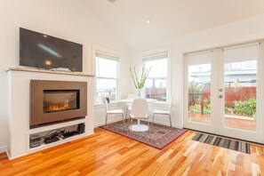 A modern living room with a fireplace, a flat-screen TV, a dining area with a white table and chairs, and a view of a garden through large sliding doors.