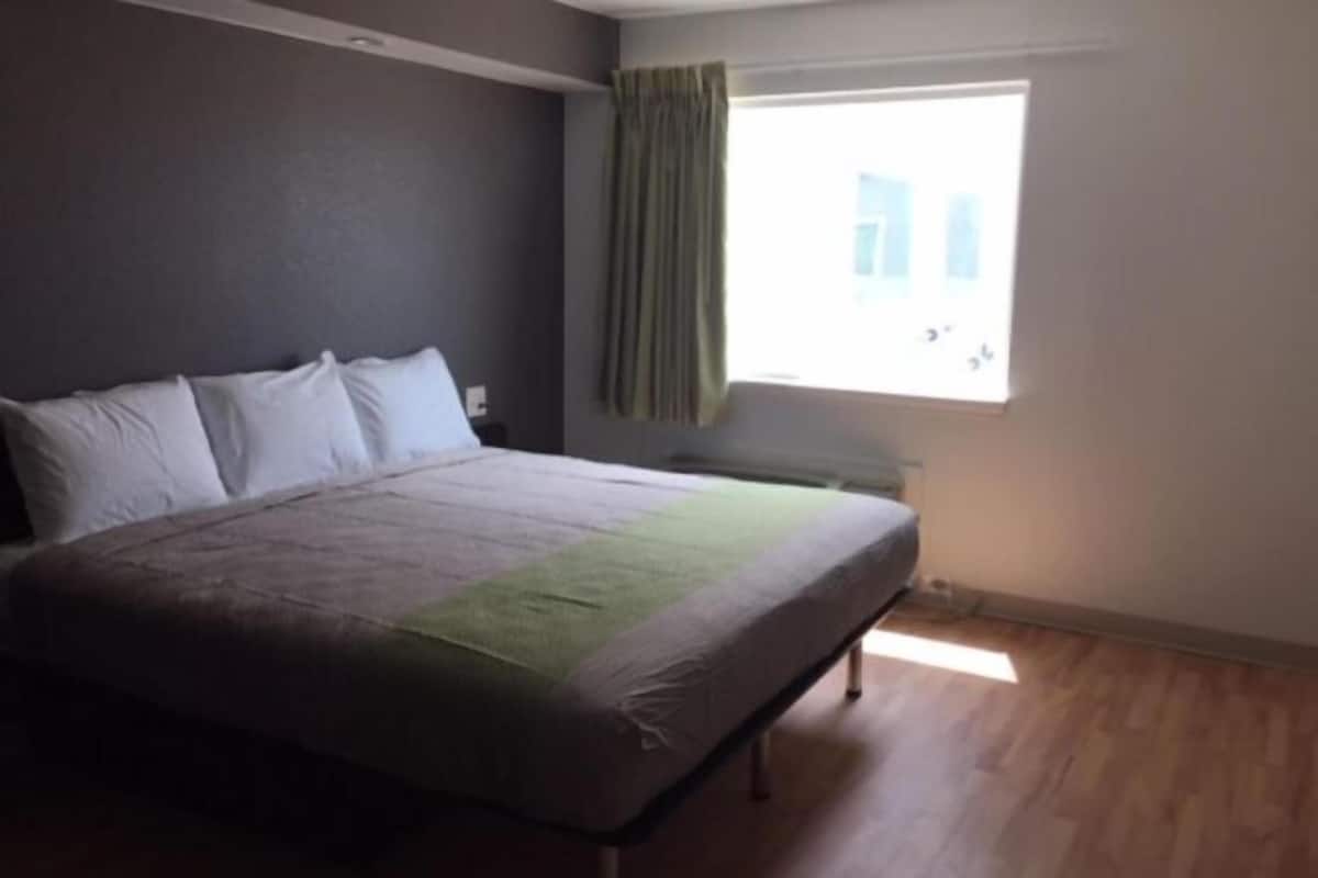 A bedroom with a bed, a chair, a window with curtains, and a view of buildings outside.