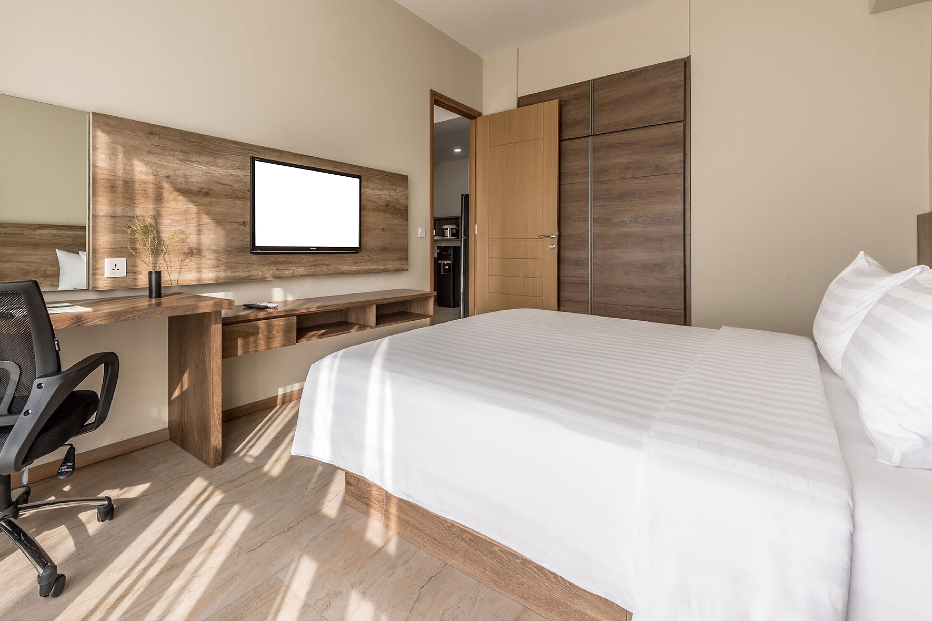 A modern hotel room with a large bed, a desk with a chair, a TV mounted on the wall, and a wardrobe.