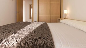 A bedroom with a bed, a wooden wardrobe, a nightstand with a lamp, and a door leading to another room. - Apartment Goleta by Interhome (Cambrils)