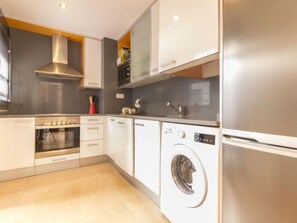A modern kitchen with a stainless steel refrigerator, built-in oven, washing machine, and a grey backsplash.