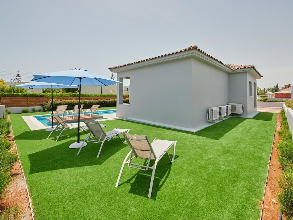 Un área de piscina con césped artificial, un pequeño edificio blanco y una sombrilla azul.