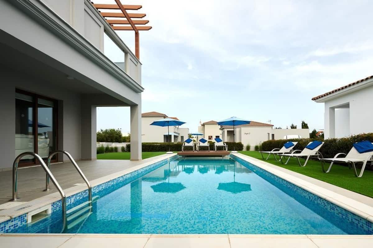 Ein moderner Pool mit klarem, blauem Wasser, umgeben von Liegestühlen und Sonnenschirmen, und ein Gebäude mit einer Pergola.