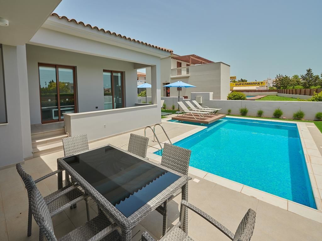 A modern outdoor pool area with a glass table and wicker chairs, surrounded by a white building with large windows.