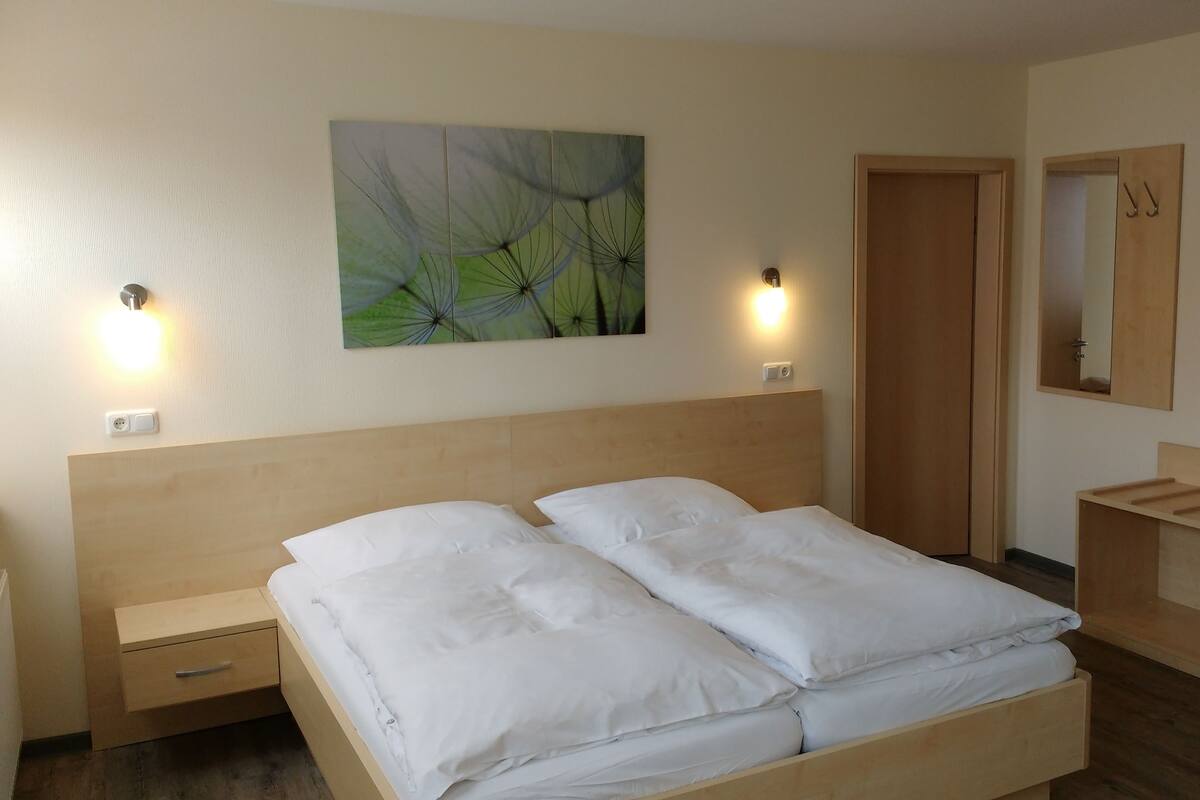 Ein ordentlich bezogenes Bett mit weißen Leinen, einem Holz-Kopfteil und einem an der Wand hängenden Gemälde mit abstrakten grünen Mustern.