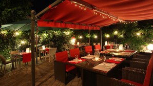 Un espace de restauration en plein air, avec des tables et des chaises, abrité par un auvent rouge et entouré de verdure.