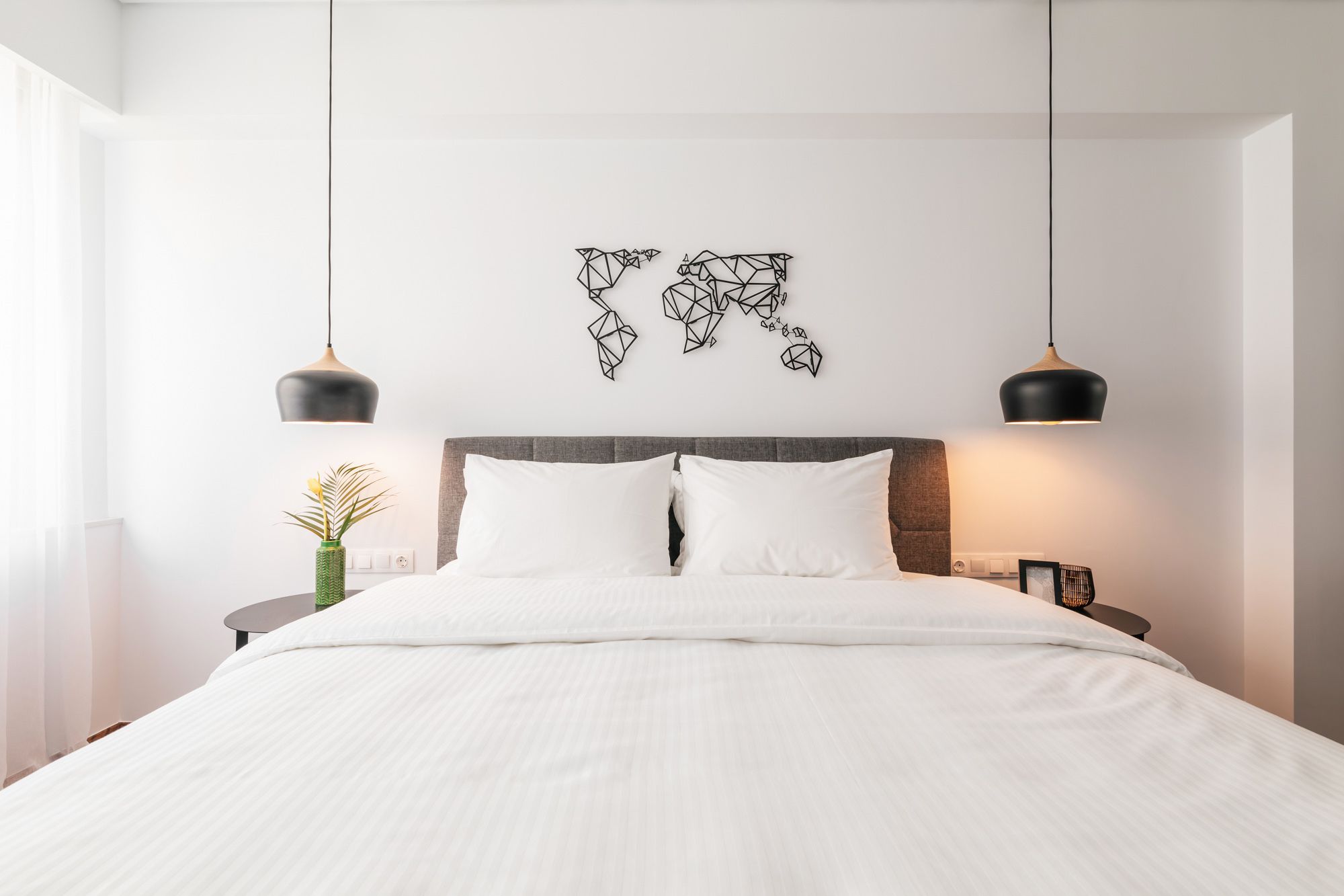 A modern bedroom with a large bed, two pendant lights, a geometric wall art piece, and a small side table with a plant.