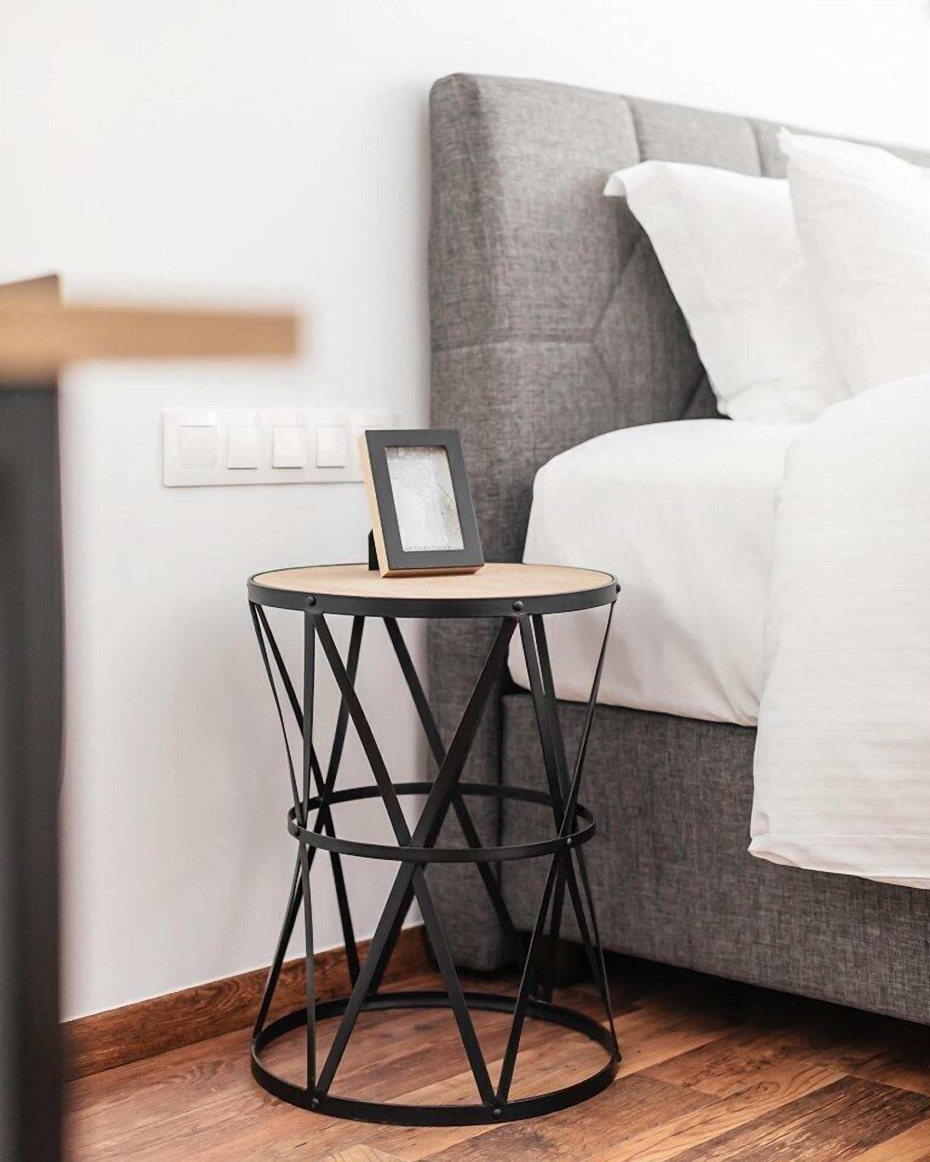 A small round side table with a black metal frame and a picture frame on it, placed next to a bed with white bedding and gray pillows.