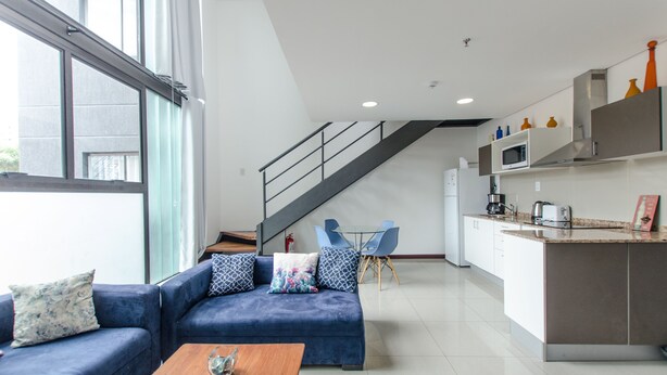 A modern living room with a blue sofa, a wooden coffee table, and a staircase leading to a kitchen area. - PrimeTime Depto en Las Mercedes (Asunción)