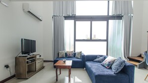 A modern living room with a blue sofa, a wooden coffee table, a TV on a wooden stand, and a large window with curtains. - PrimeTime Depto en Las Mercedes (Asunción)