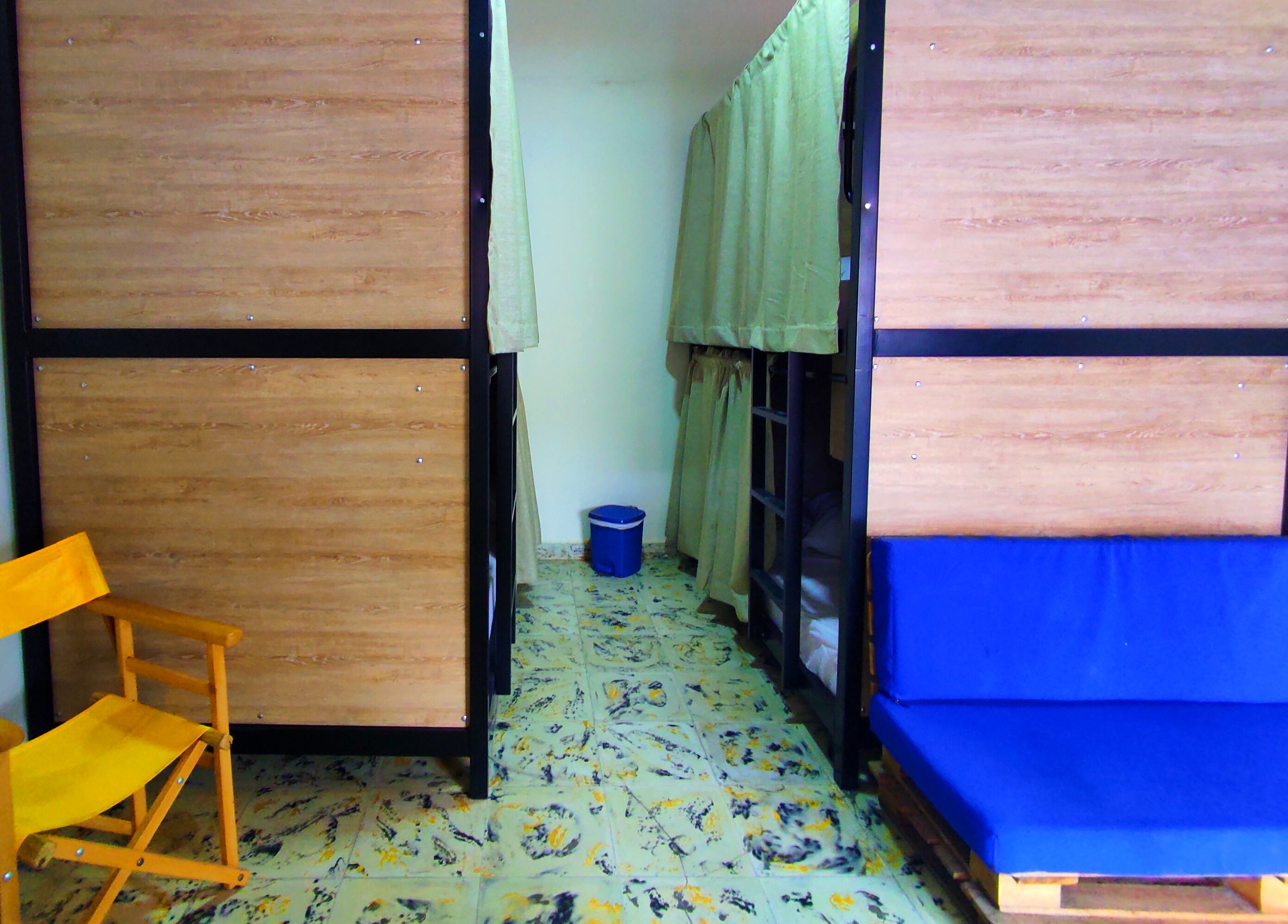 A small, compact room with a wooden bunk bed, a foldable chair, and a blue trash bin.
