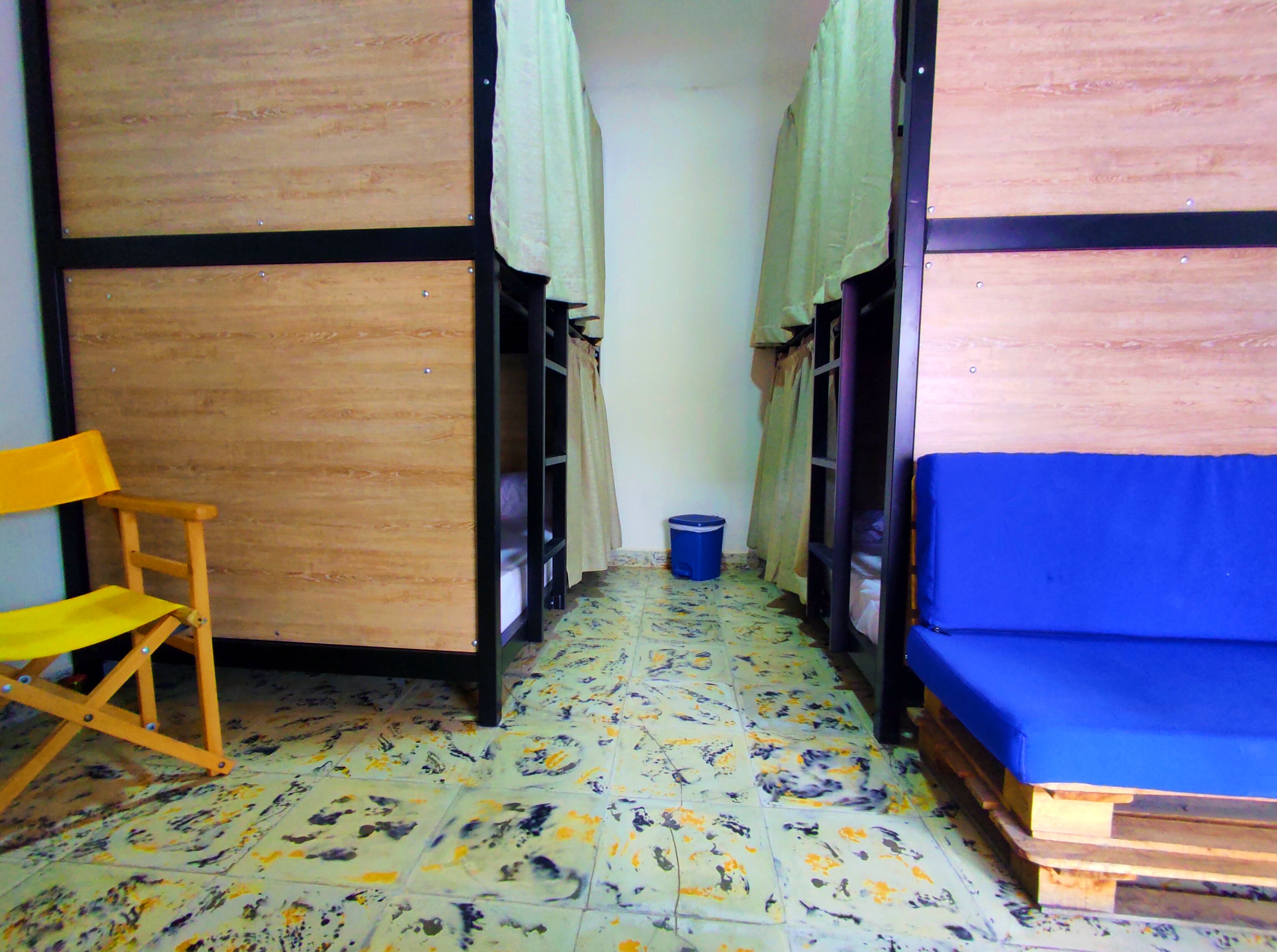 Un estrecho pasillo con literas a ambos lados, una silla de madera y un colchón azul a la derecha.