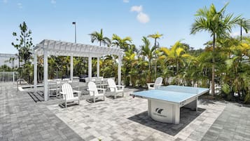 Un espace extérieur aménagé avec une table de ping-pong, des chaises blanches et une structure abritée pour s’asseoir.