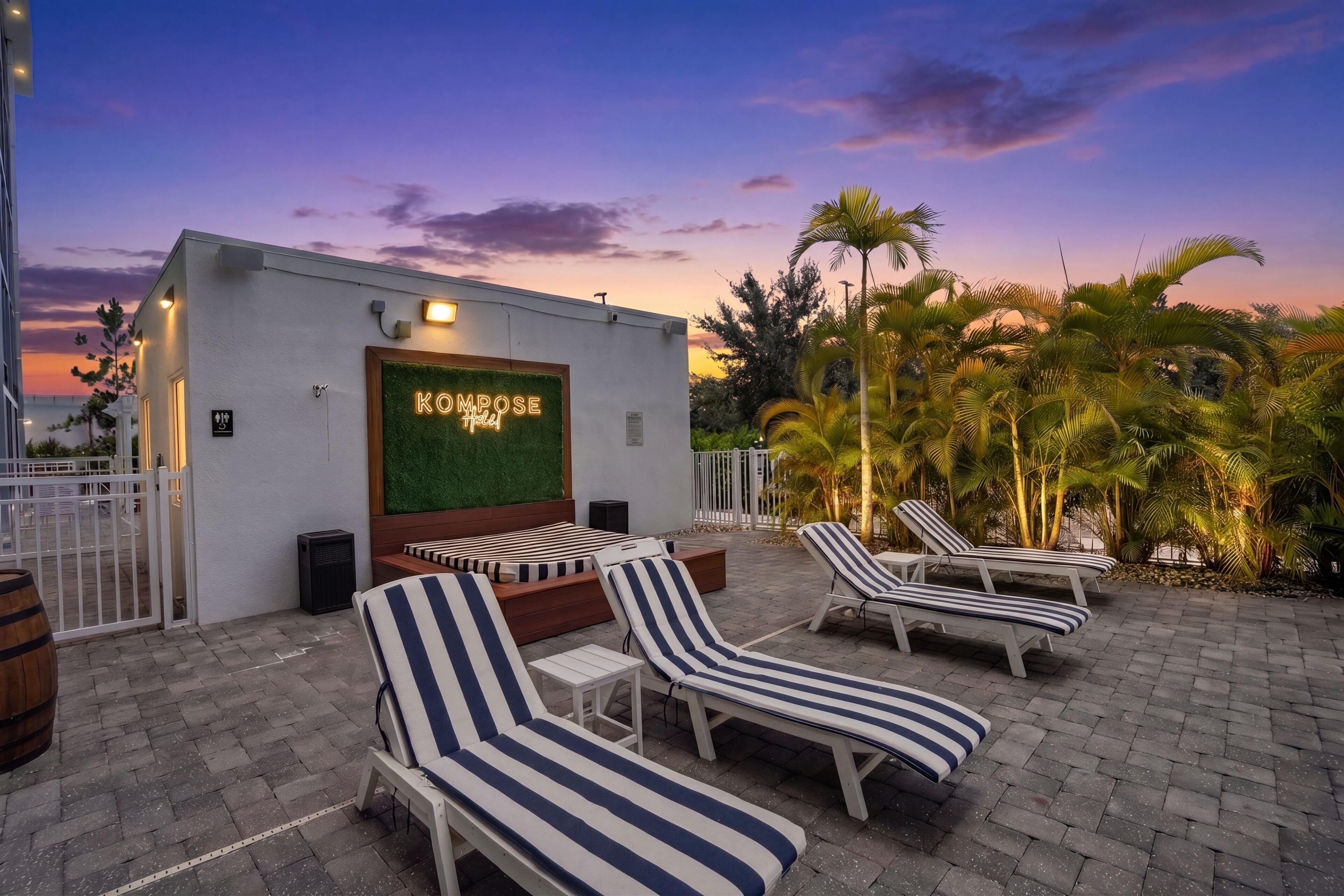 A rooftop lounge area with striped lounge chairs, a neon sign reading 'KOMPOSE Hotel', and a view of the sunset.