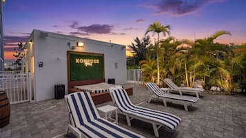 A rooftop lounge area with striped lounge chairs, a neon sign reading 'KOMPOSE Hotel', and a view of the sunset.