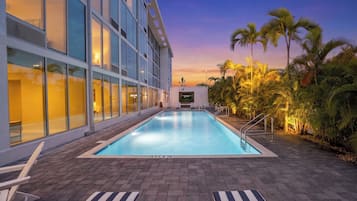 A swimming pool with lounge chairs, a building with large windows, and palm trees.