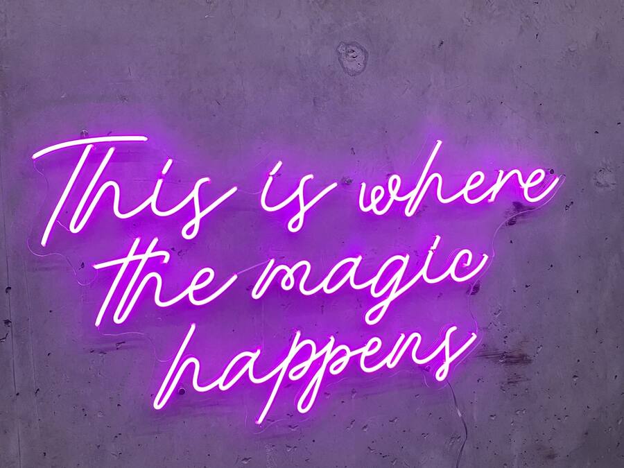 A neon sign on a textured wall with the text "This is where the magic happens."