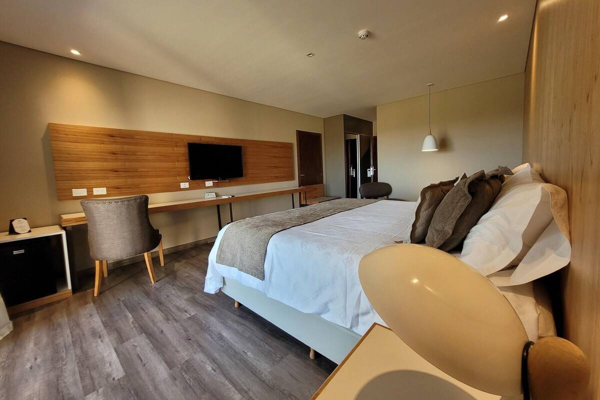 A modern hotel room with a large bed, a desk with a chair, a television, and a wardrobe.