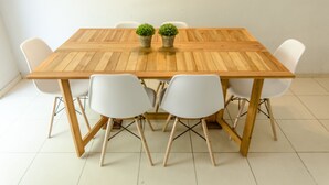 A rectangular wooden table with white chairs, two potted plants, and a white wall in the background.
