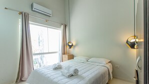 A neatly made bed with rolled towels, a bedside table, a wall-mounted air conditioner, and two wall sconces.