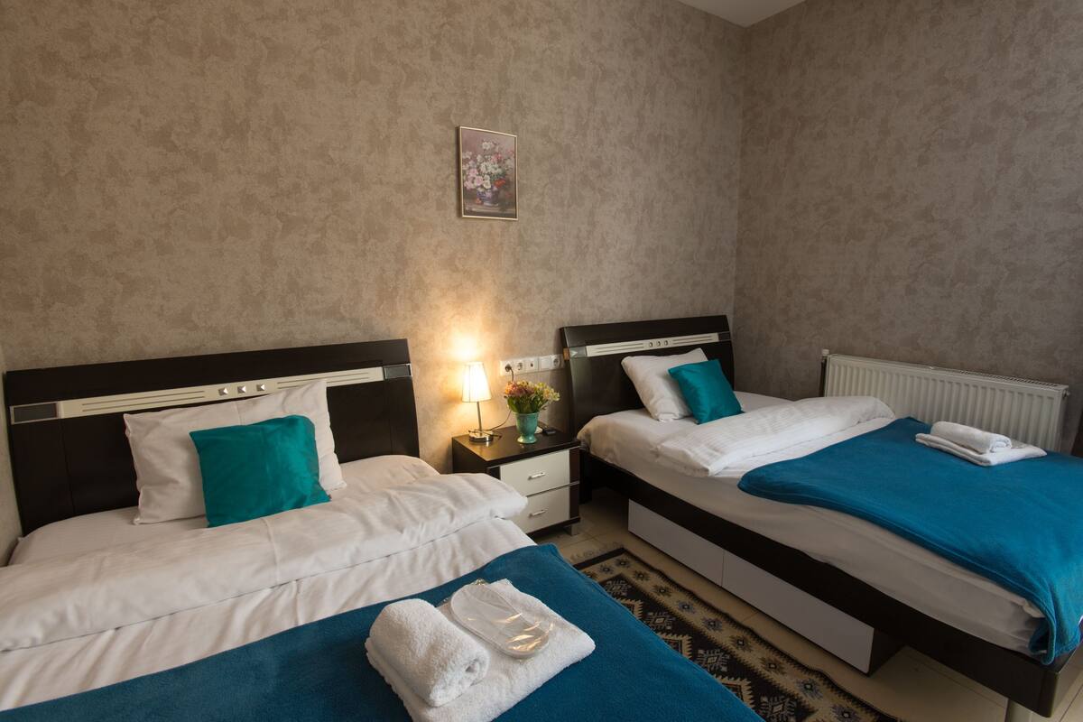 A hotel room with two beds, a nightstand with a lamp, and a radiator.