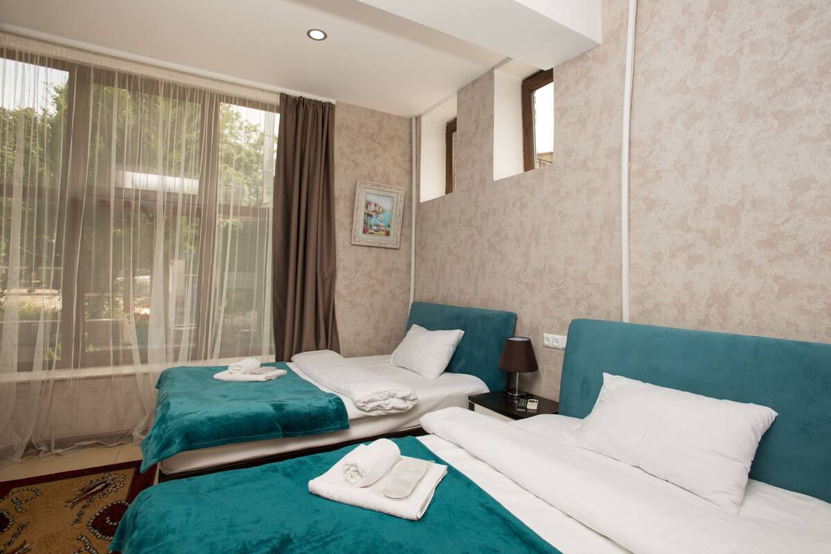 A hotel room with two beds, a large window with sheer curtains, a painting on the wall, and a small bedside table with a lamp.