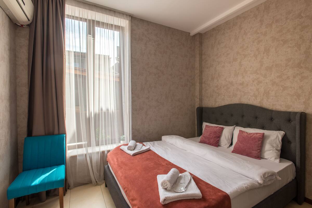 A hotel room with a bed, a blue chair, and a window with sheer curtains.