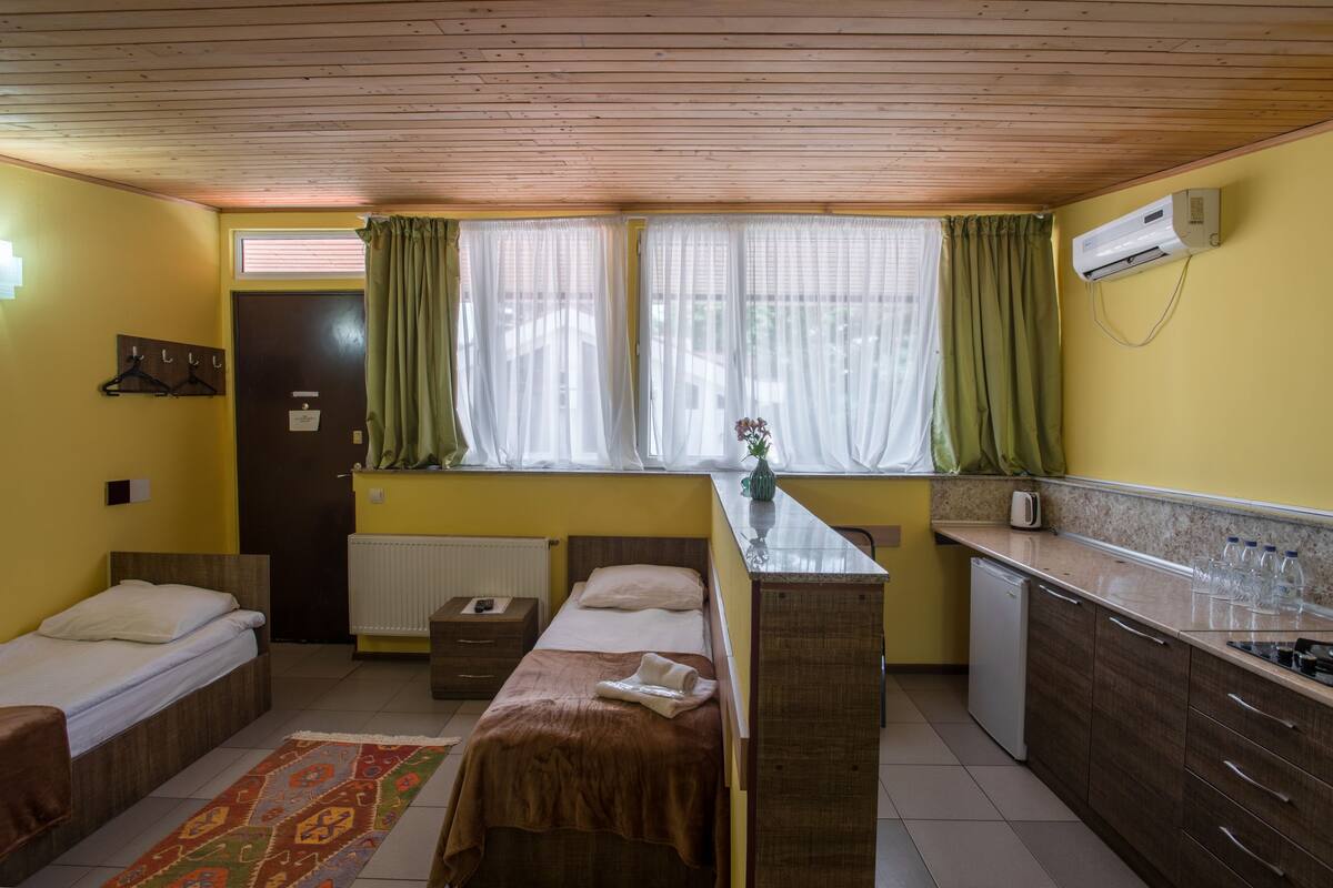 A compact room with two beds, a small kitchenette, a radiator, and a window with curtains.