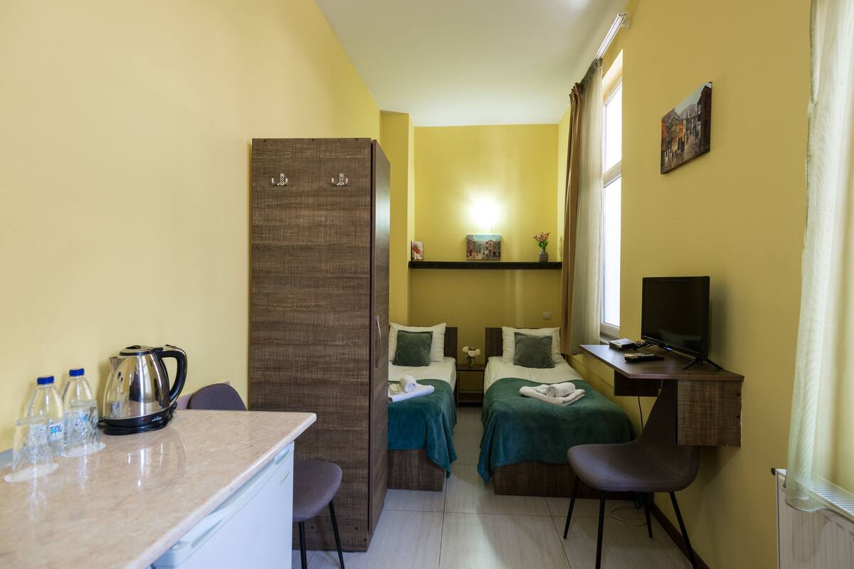 A compact hotel room with two beds, a desk with a computer, a small kitchenette, and a window with curtains.
