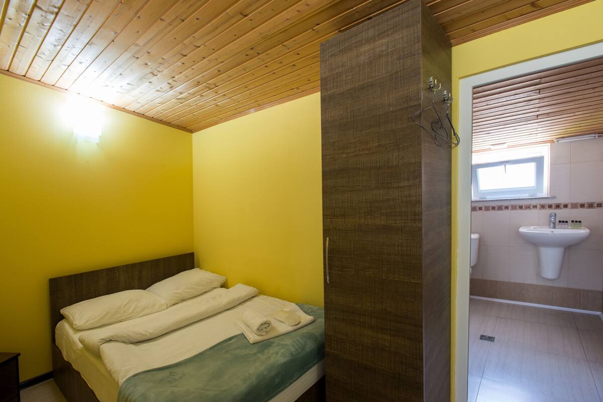 A room with a bed, a wooden ceiling, and a bathroom with a sink and window.
