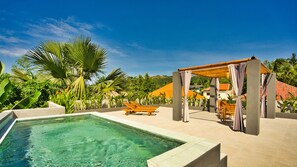 A pool area with a clear blue pool, lounge chairs, and a pergola with curtains. - Dalai Home (Kuta)