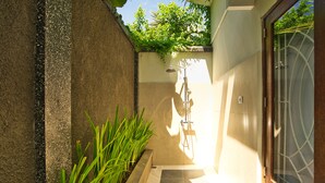 A modern shower area with a glass door, a tiled floor, and a small planter with green plants. - Dalai Home (Kuta)