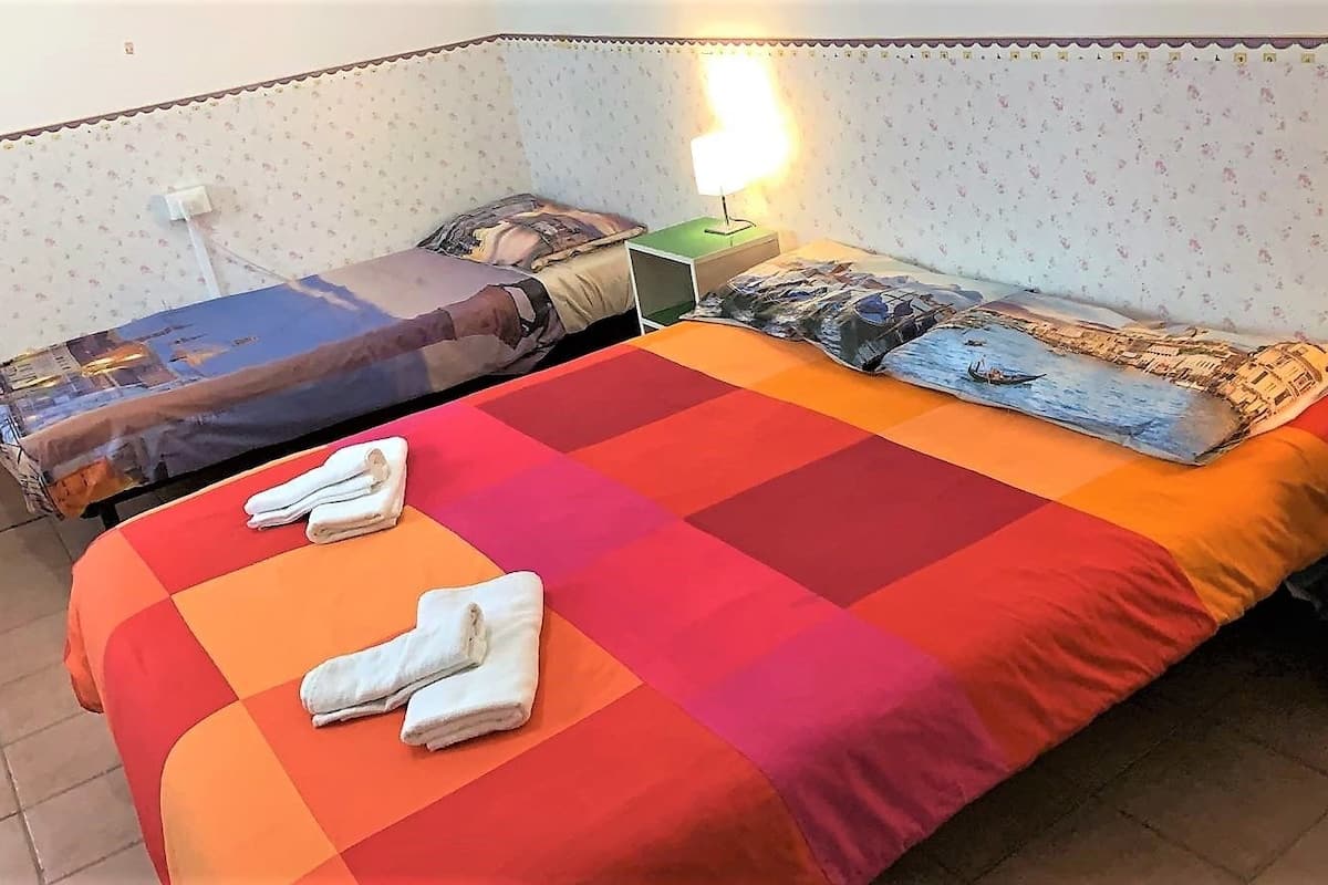 A room with two beds, each with a different patterned bedspread. There are two bedside tables with lamps, and towels are neatly folded on the beds.