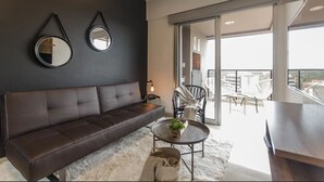 A modern living room with a dark brown sofa, two wall-mounted mirrors, a round coffee table with a plant, and a view of the city through large windows. - Modern 1 BR Apartment (Asunción)