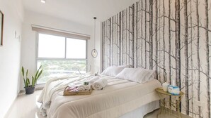 A modern bedroom with a large window, a bed with white bedding, a small round table, and a wall with a tree pattern. - Modern 1 BR Apartment (Asunción)